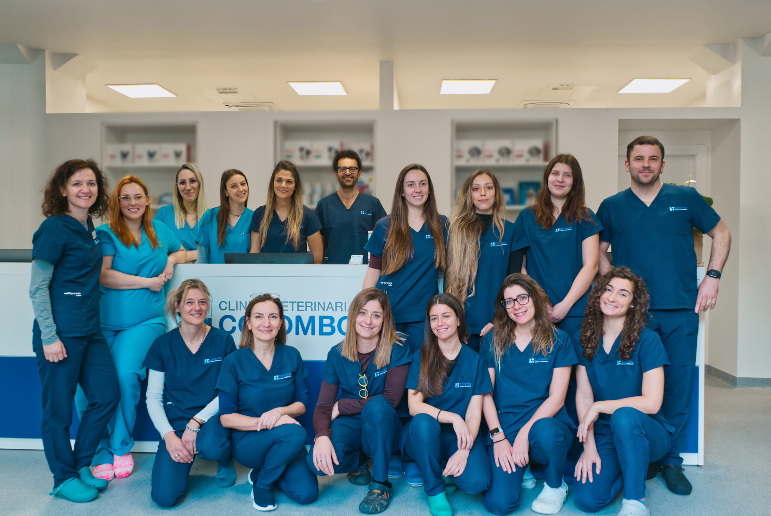 L'immagine ritrae *Il team* di veterinari e personale in camice blu, sorridenti e in posa insieme nella reception di una clinica veterinaria. Sullo sfondo sono visibili un bancone della reception e prodotti per la cura degli animali.