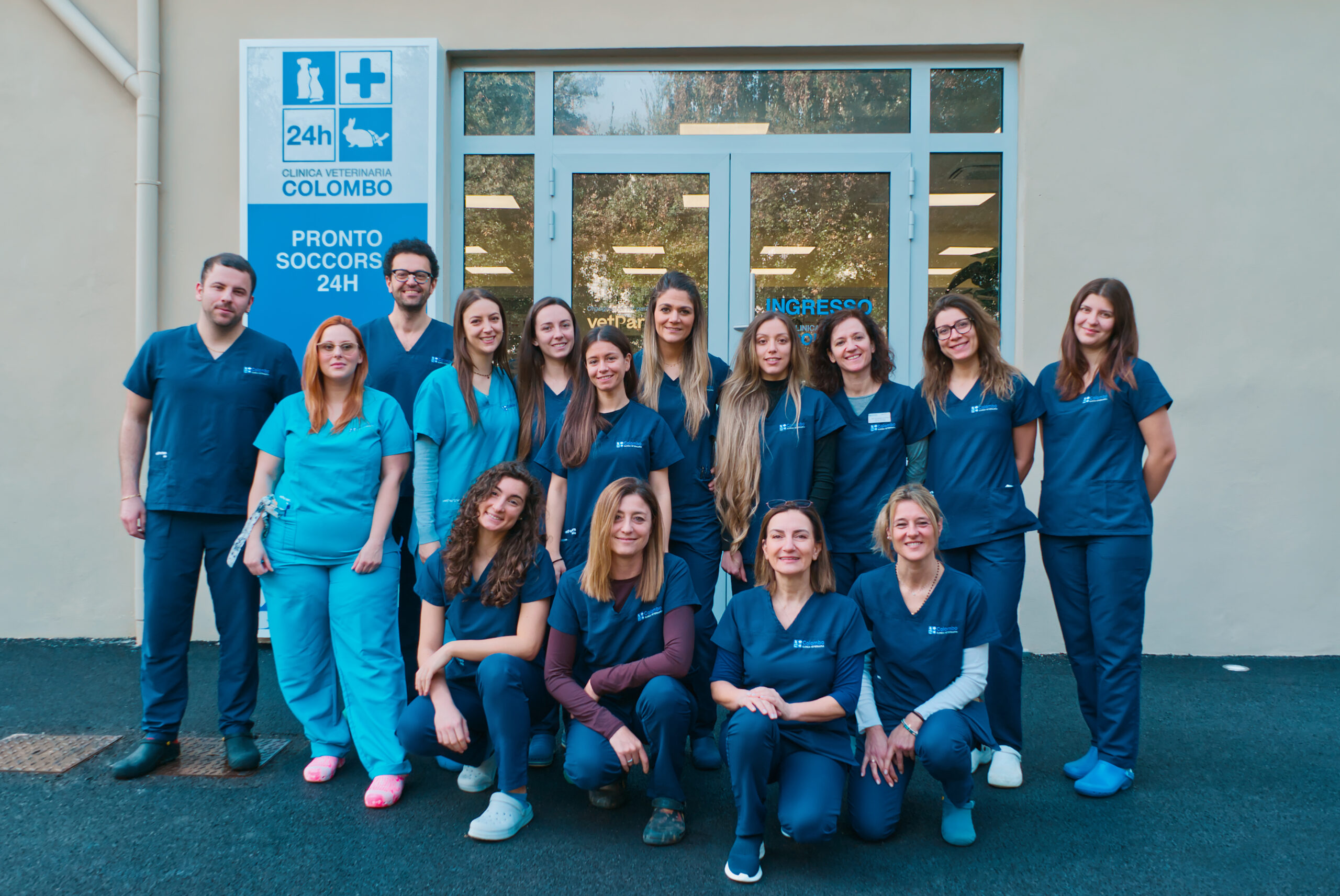 Il personale dedicato della Clinica Veterinaria Colombo si erge fiero all'esterno, vestito con uniformi blu. Accanto a loro, un cartello decorato con simboli di animali domestici annuncia servizi di emergenza 24 ore su 24. Le porte e le finestre in vetro della clinica riflettono il loro approccio professionale ma amichevole.