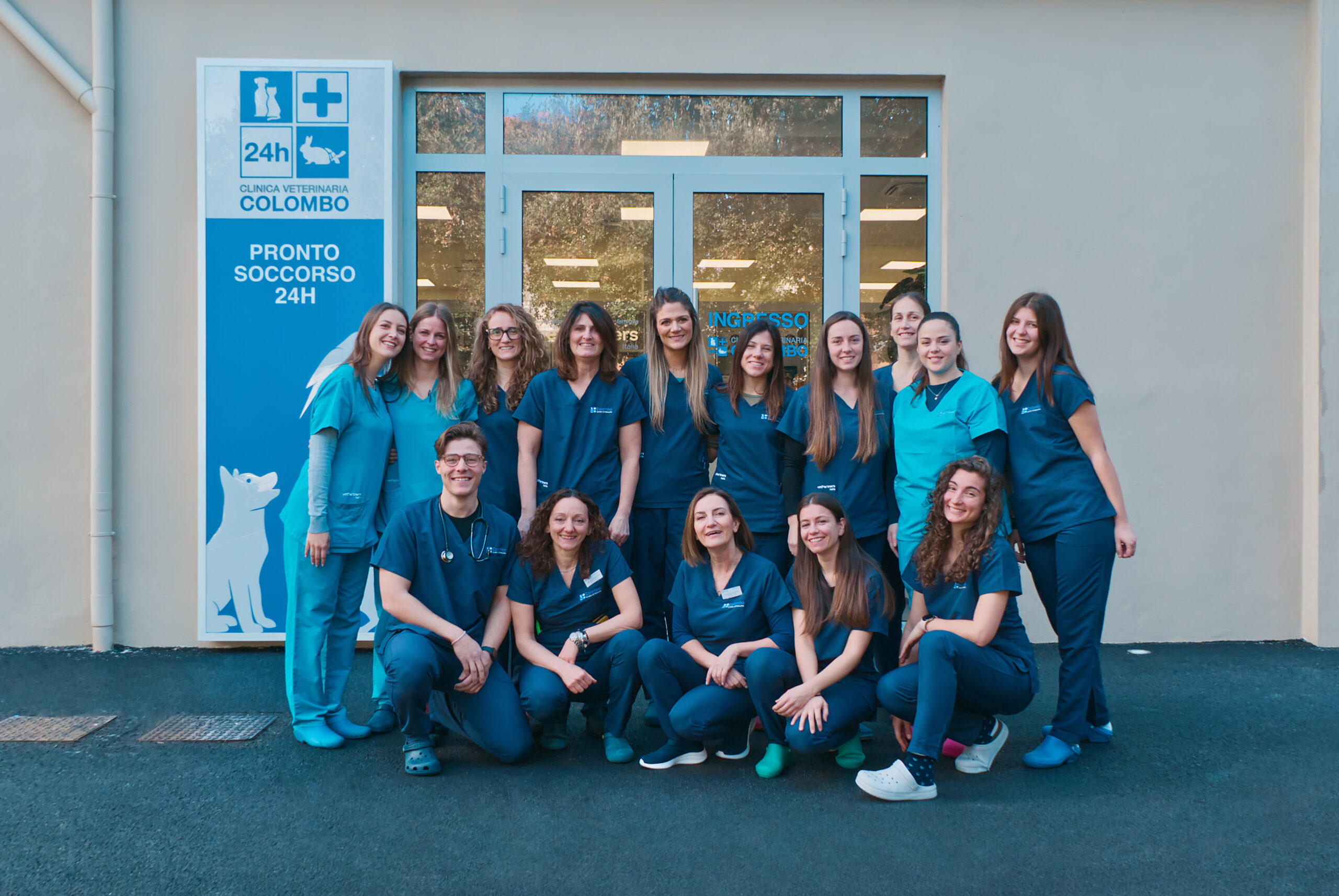 Un gruppo di sedici persone in camice blu posa all'ingresso della Clinica Veterinaria Colombo. Accanto a loro, un cartello con un disegno di un gatto e un cane recita "24h Pronto Soccorso", evidenziando i loro servizi di emergenza 24 ore su 24.