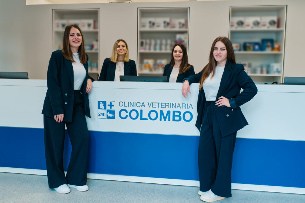 Un team di quattro donne in abiti da lavoro è in piedi e seduto attorno a una reception di una clinica veterinaria. La reception espone con orgoglio un cartello, "Clinica Veterinaria Colombo", decorato con simboli medici, mentre sullo sfondo si vedono scaffali pieni di prodotti per animali domestici.