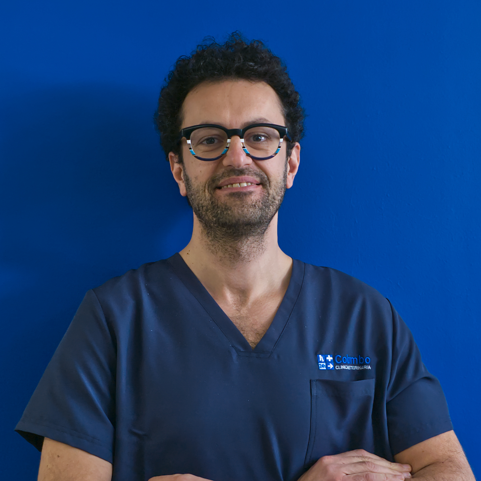 Una persona con i capelli ricci e gli occhiali sorride mentre indossa un camice medico blu, in piedi su uno sfondo blu uniforme. Irradia la sicurezza e la compassione del team, incarnando sia la professionalità che il calore nel loro impegno costante per la cura.