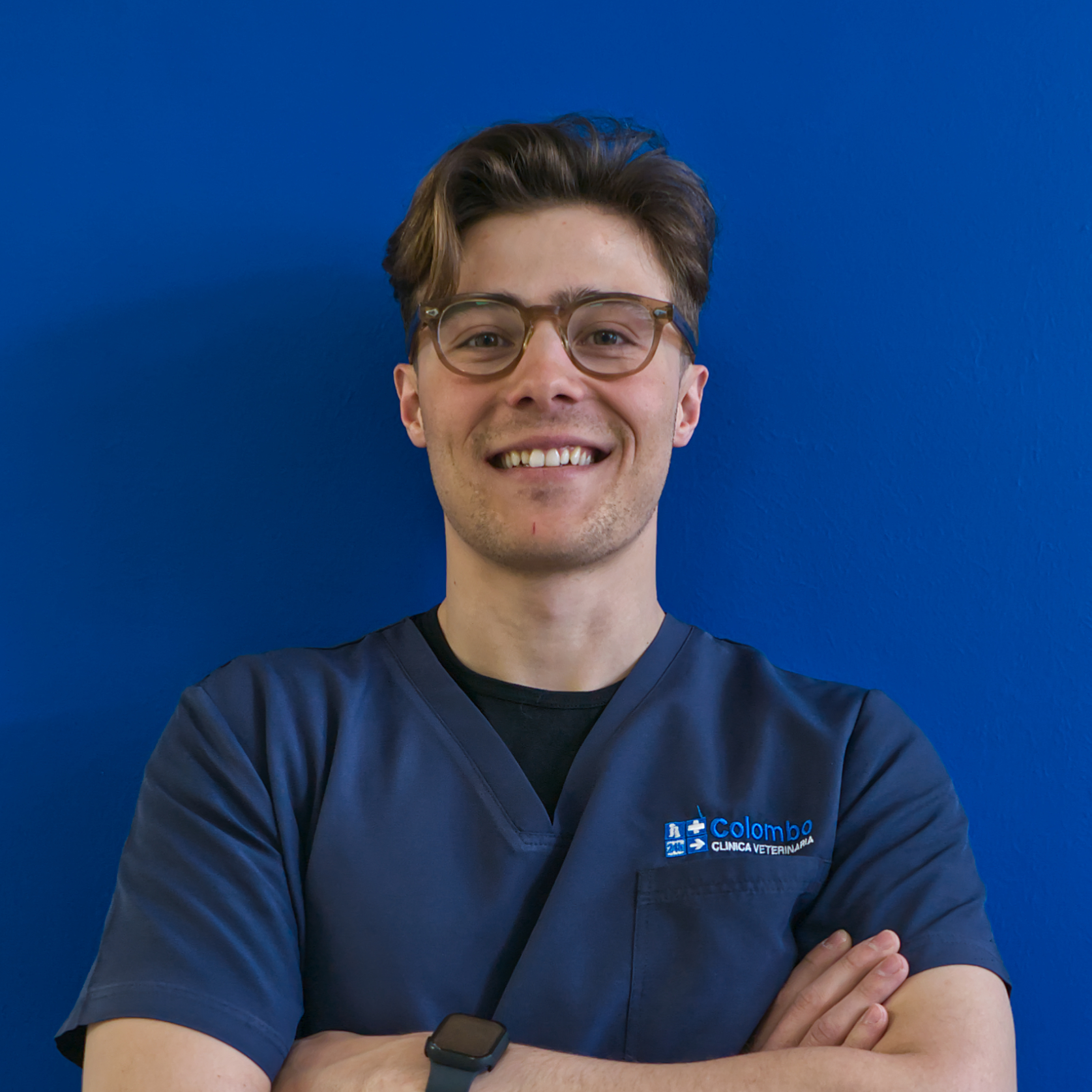 Un membro del team, con i capelli corti castani e gli occhiali, sorride con sicurezza, con le braccia incrociate, in una camicetta blu navy su uno sfondo blu brillante.