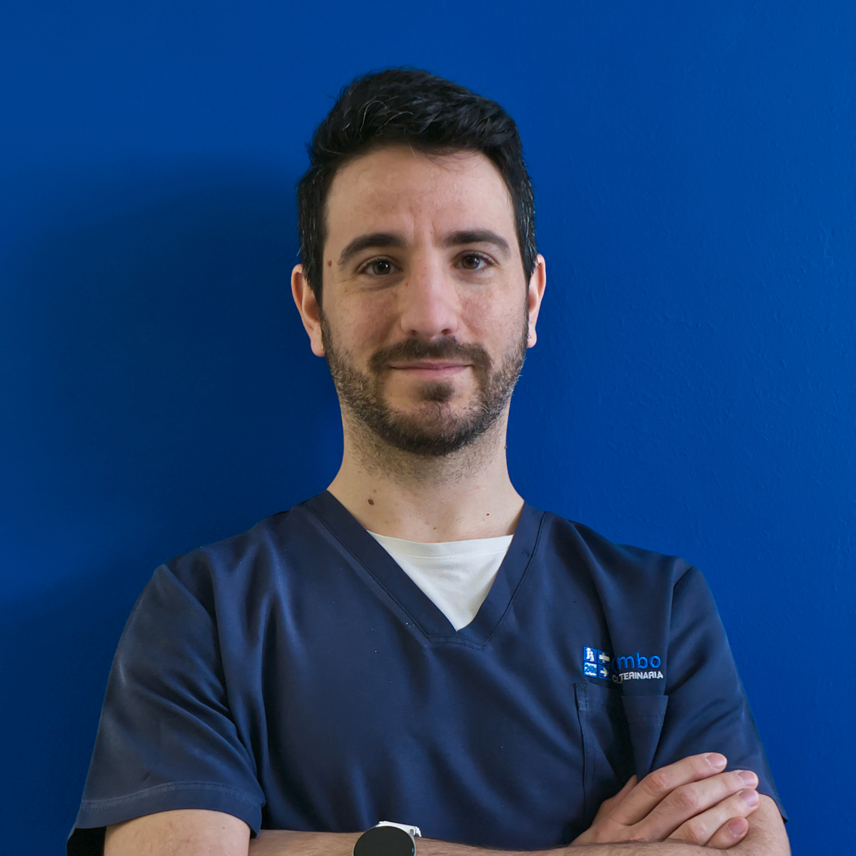 Contro una parete blu, un uomo con i capelli scuri e la barba curata sta in piedi con sicurezza. Indossando una camicetta blu navy sopra una canottiera bianca, con le braccia incrociate, incarna la professionalità del team mentre guarda la telecamera con un'espressione neutra.