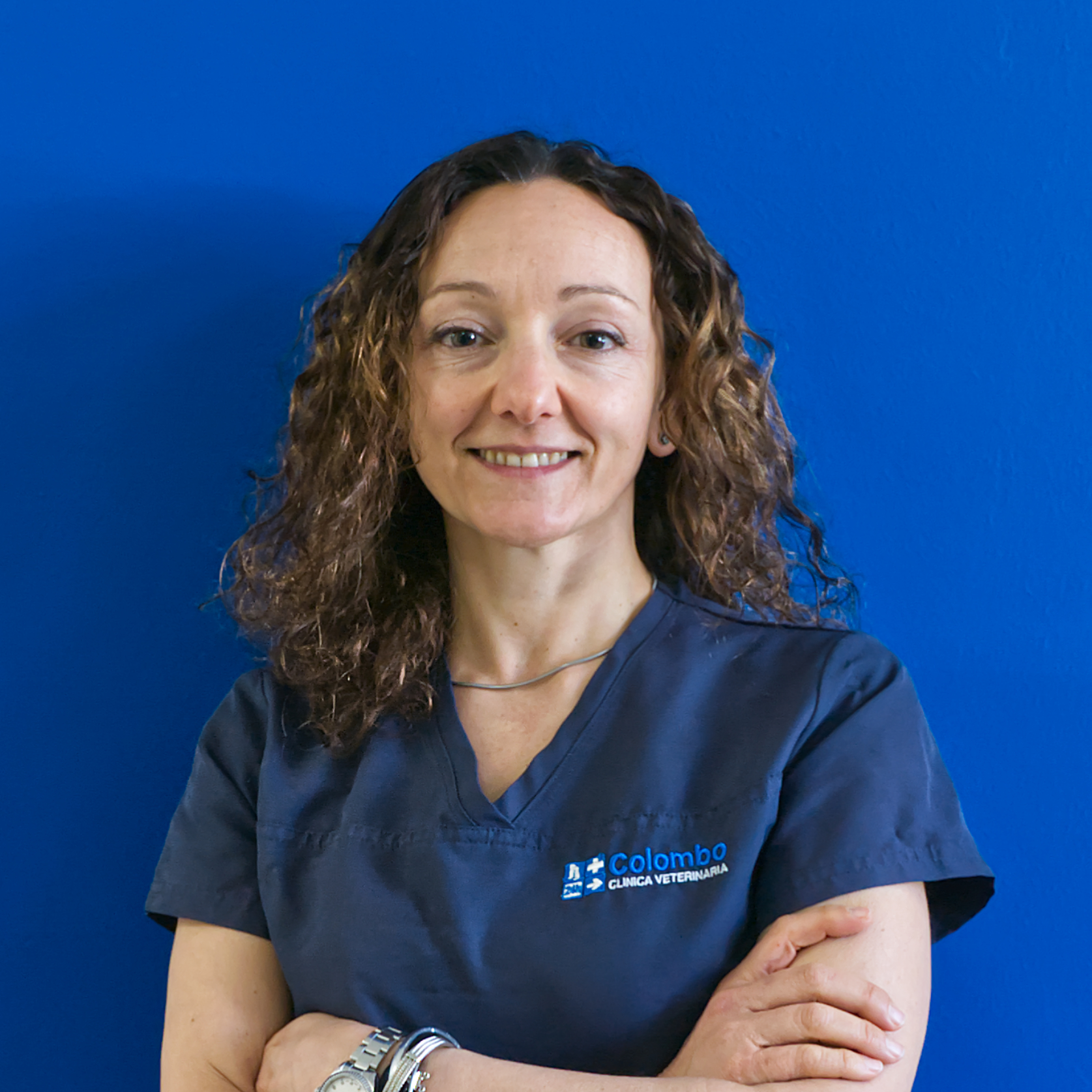 Un membro del team, una donna con i capelli ricci e un camice blu, sta in piedi con sicurezza, con le braccia incrociate, e sorride calorosamente davanti a un muro blu brillante.