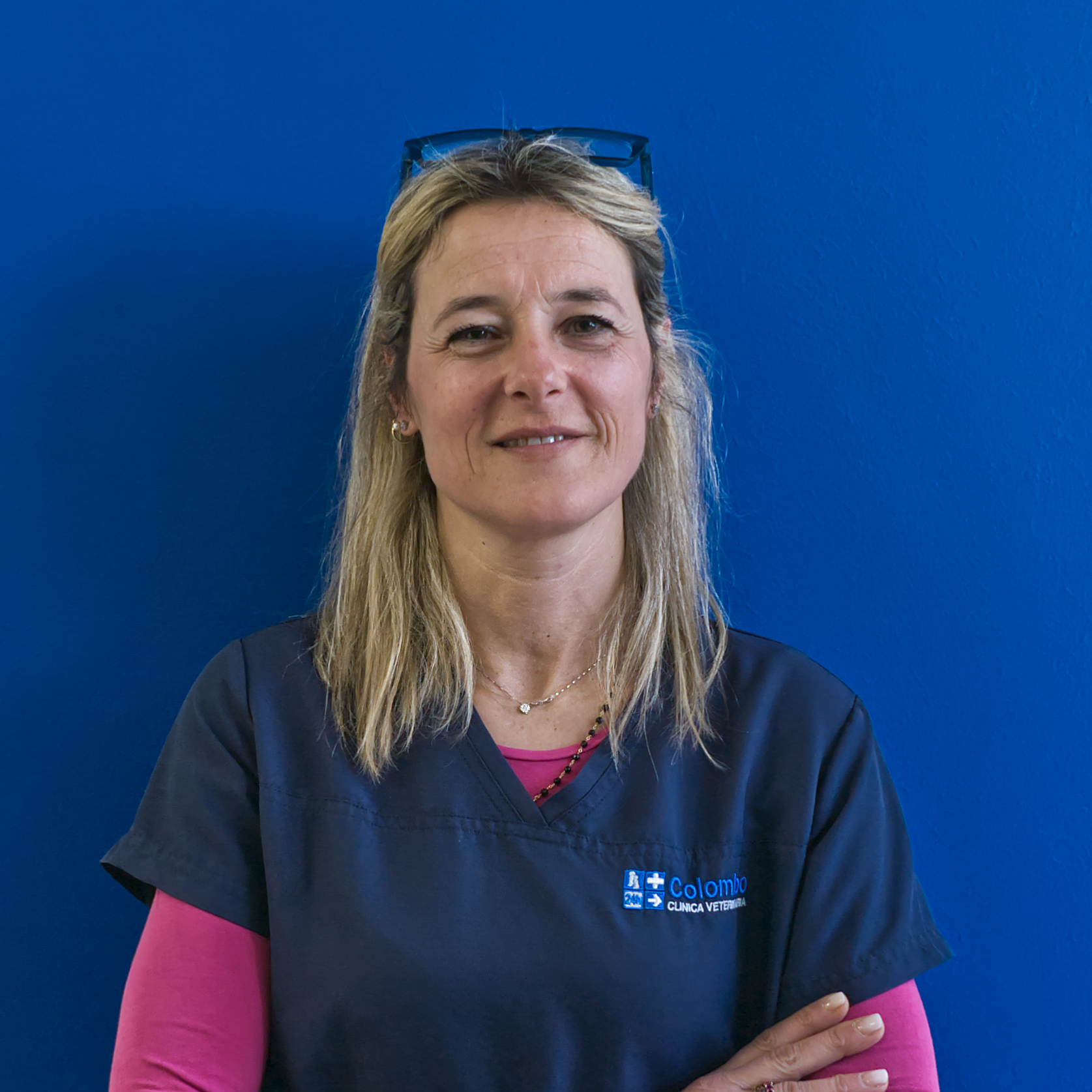 Un membro del team con lunghi capelli biondi sta in piedi su uno sfondo blu brillante, indossando un camice blu navy decorato con un logo su una maglietta rosa. Indossa occhiali da sole in testa, incrocia le braccia e sorride leggermente.