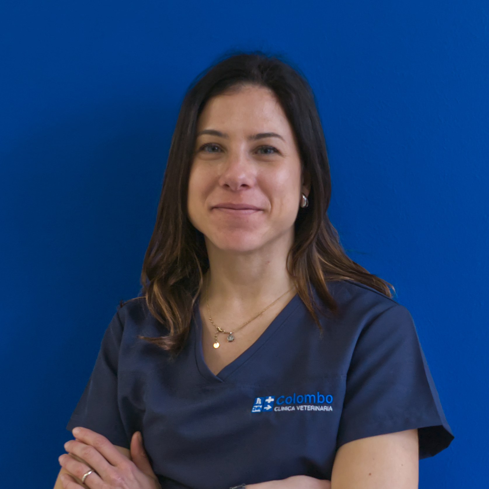 Una donna con lunghi capelli scuri è in piedi con sicurezza, con le braccia incrociate, e sorride leggermente. Indossa con orgoglio una maglietta blu navy decorata con il logo della squadra Il. Lo sfondo è un muro blu uniforme, che aggiunge unità e professionalità alla scena.