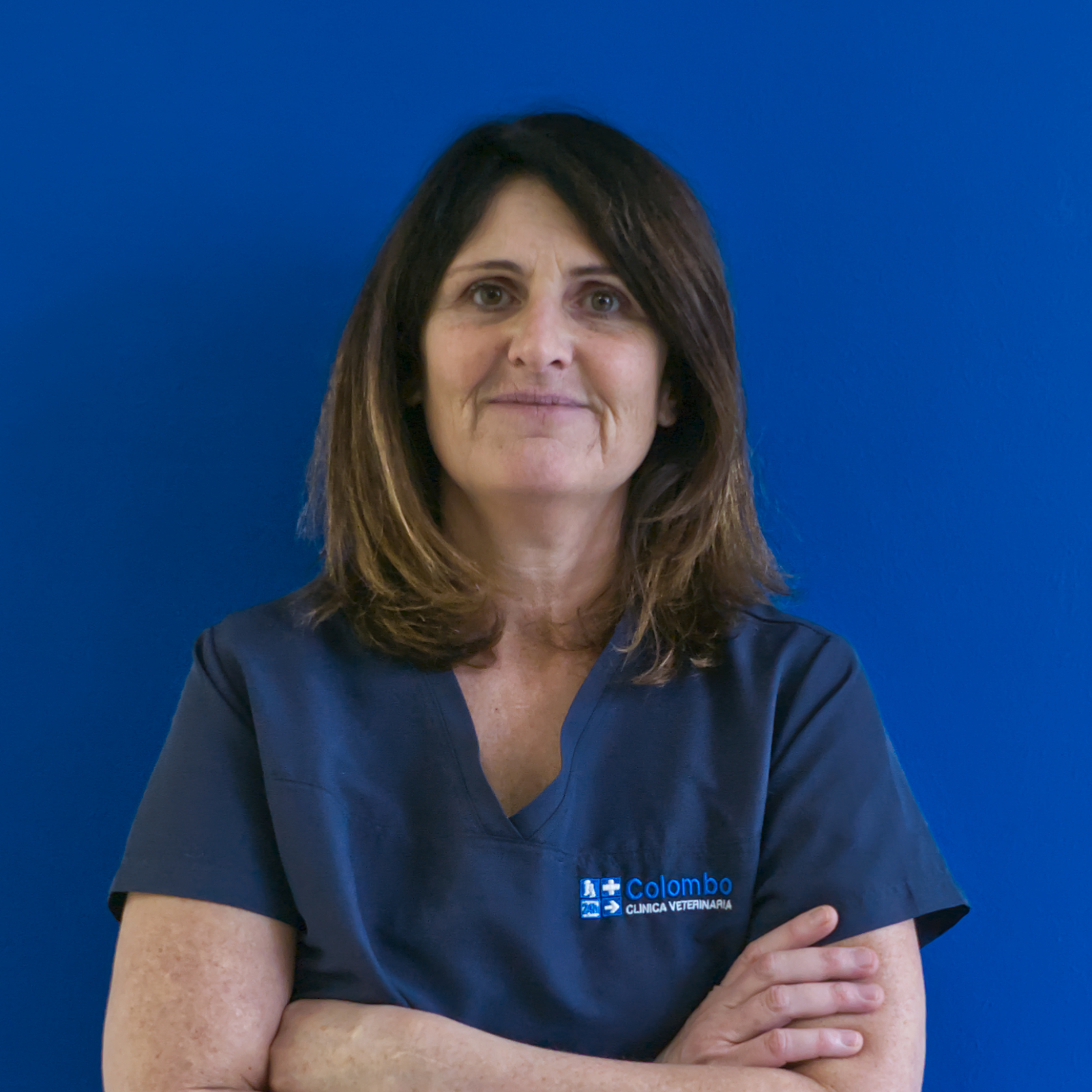 Una donna con i capelli castani lunghi fino alle spalle è in piedi su uno sfondo blu, con la camicetta blu navy ricamata con la scritta 