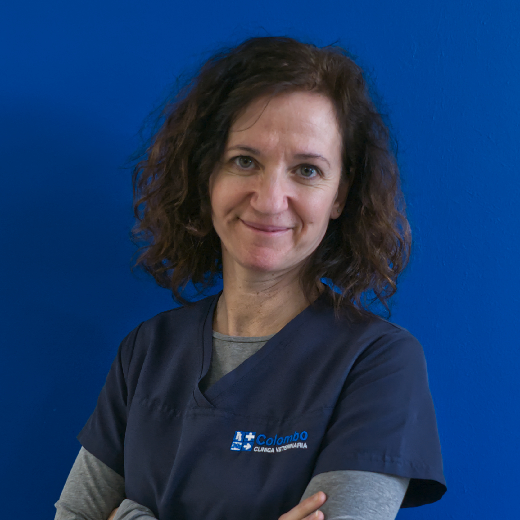 Una donna con i capelli scuri e ricci sorride su uno sfondo blu. Indossa un camice blu navy con un logo, che rappresenta il suo ruolo nella squadra, e ha le braccia incrociate con sicurezza.
