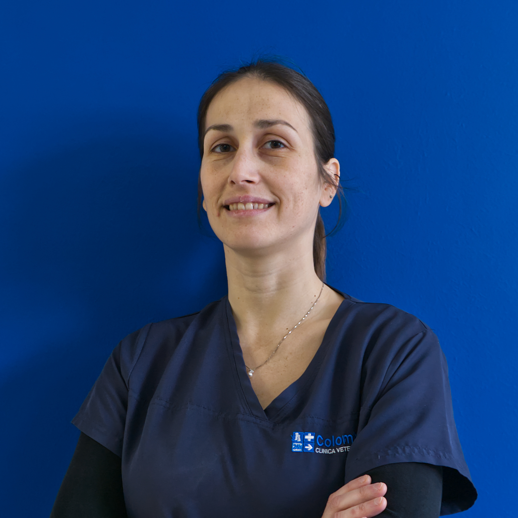Una donna in camice blu navy è in piedi con sicurezza, con le braccia incrociate, contro un muro blu brillante; il suo leggero sorriso riflette lo spirito del 
