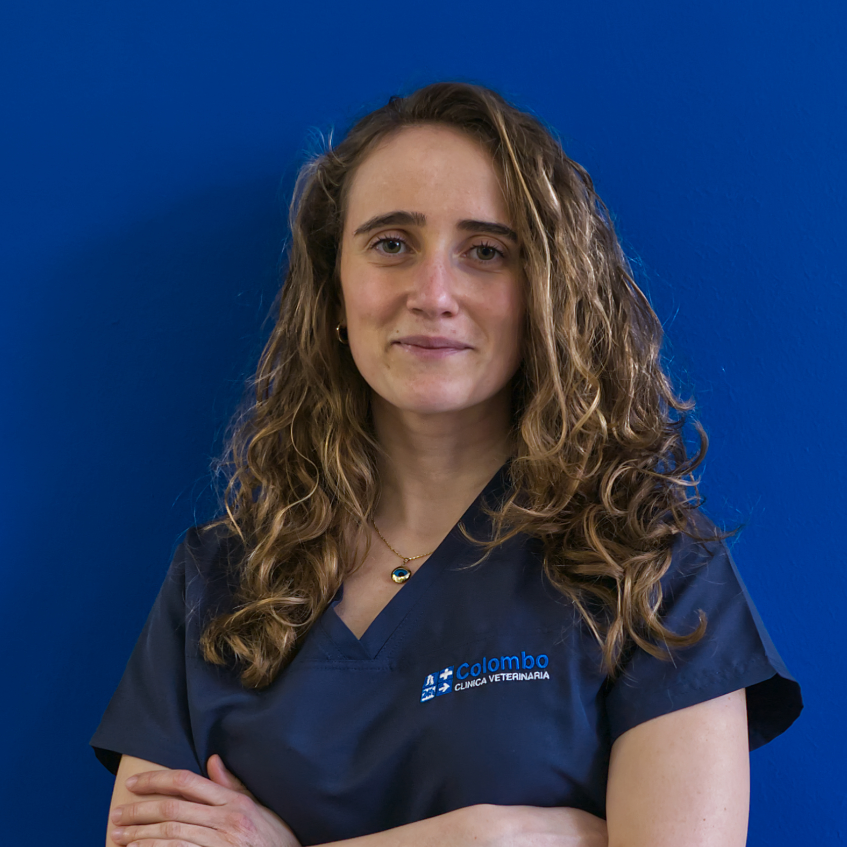 Una donna con lunghi capelli ricci sta in piedi con sicurezza su uno sfondo blu, indossando un camice blu navy con un logo sul petto. Rappresenta la squadra con le braccia incrociate e un sorriso gentile, guardando dritto in camera.