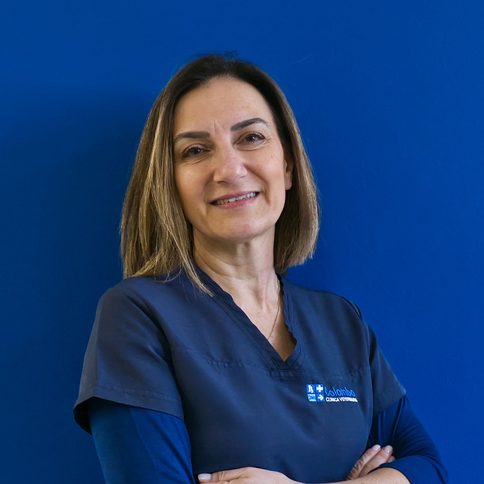 Una donna con i capelli castani lunghi fino alle spalle, che indossa una camicetta blu navy, sorride con sicurezza con le braccia incrociate su uno sfondo blu uniforme. È un membro chiave del team, incarnando lo spirito di professionalità e dedizione.