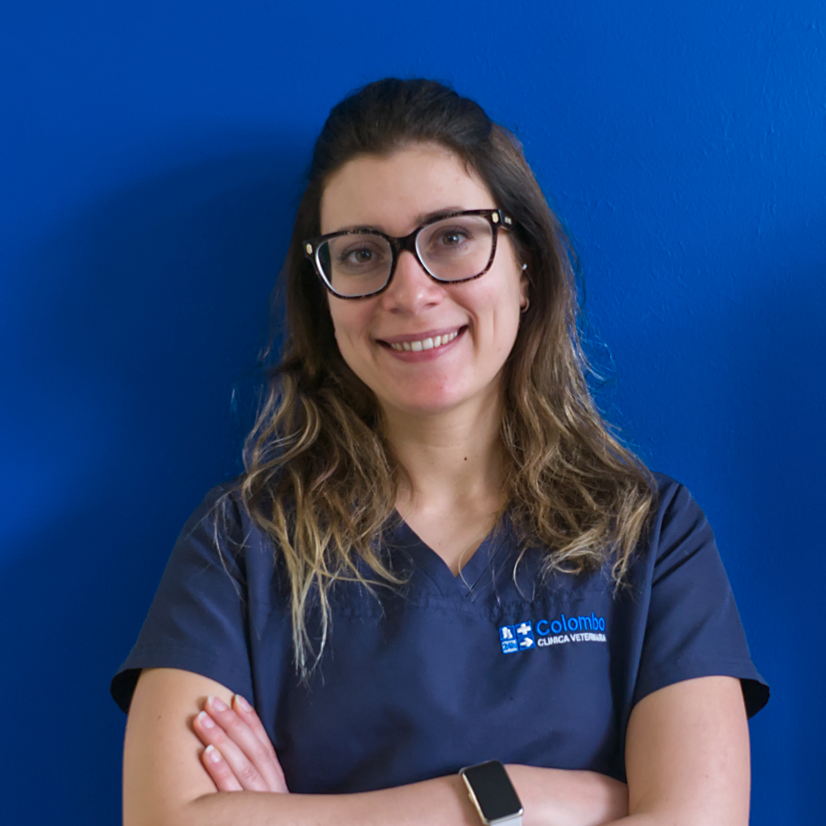 Una donna con lunghi capelli ondulati e occhiali sorride alla telecamera. Indossa un camice medico blu scuro e sta in piedi con sicurezza con le braccia incrociate contro un muro blu uniforme. Il suo atteggiamento accogliente riflette la professionalità e il calore del team che rappresenta con orgoglio.