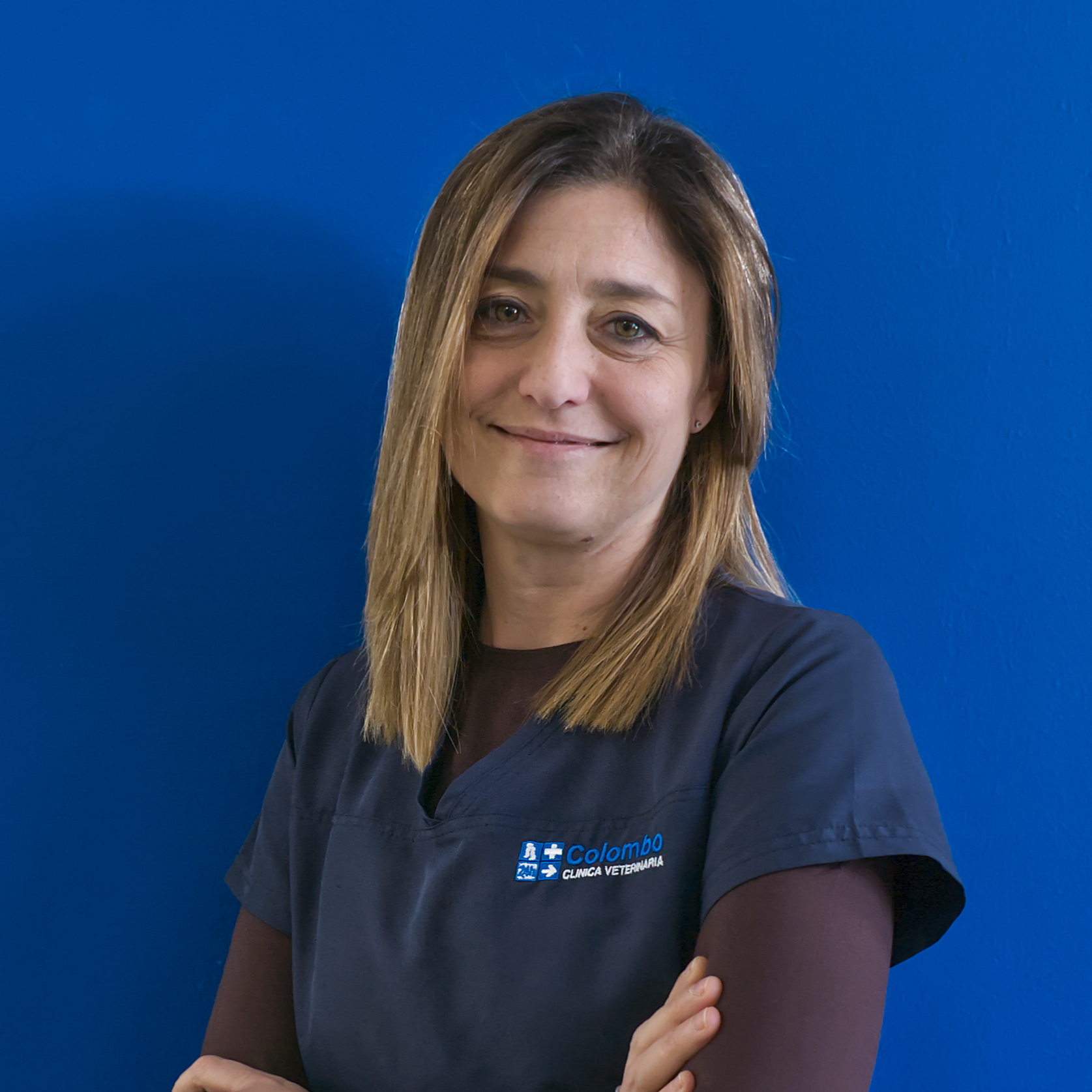 Una donna con i capelli biondi lisci e lunghi fino alle spalle sorride con sicurezza alla telecamera. Indossa un camice blu scuro con un logo, sta in piedi su uno sfondo blu brillante con le braccia incrociate, incarnando lo spirito del team il.
