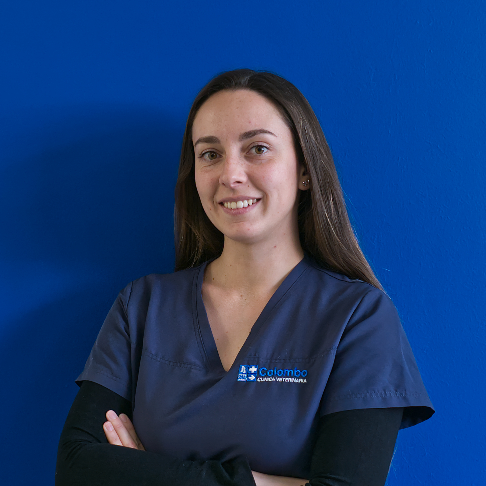 Una donna con lunghi capelli castani sorride e incrocia le braccia con sicurezza. Indossa un camice medico blu decorato con un logo sul lato sinistro, che rappresenta il team Il. Lo sfondo è di un colore blu uniforme, che esalta la sua presenza professionale.