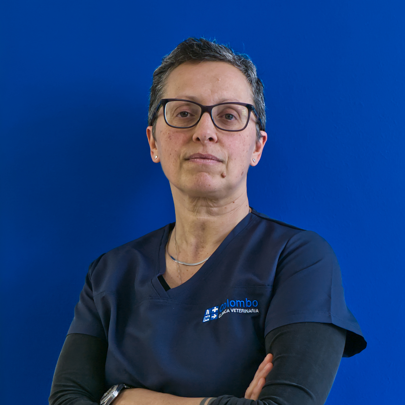 Un membro del team con i capelli corti e gli occhiali si erge con sicurezza su uno sfondo blu brillante, indossando un camice blu scuro con ricamata la scritta 