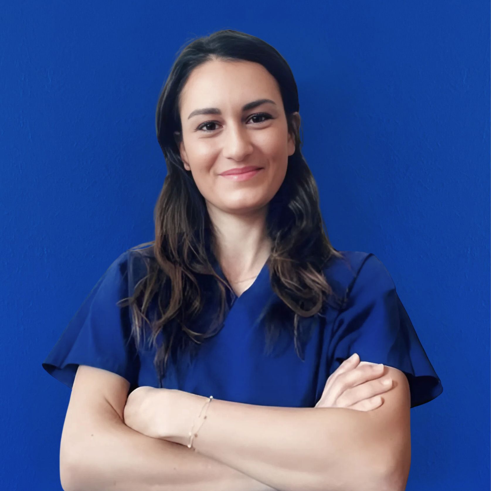 Una donna con lunghi capelli castani, che indossa un camice blu, sta in piedi con sicurezza con le braccia incrociate su uno sfondo blu. Mentre sorride dolcemente, irradia professionalità e riflette l'impegno del team che guida l'eccellenza insieme.