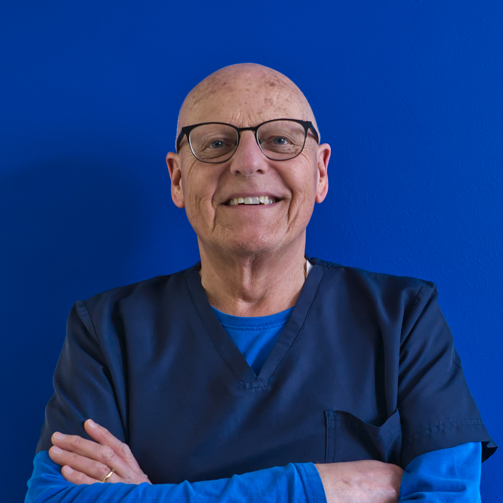 Un individuo sorridente con gli occhiali e la testa calva sta in piedi su uno sfondo blu acceso, indossando una camicetta blu sopra una camicia a maniche lunghe. Lo spirito di squadra traspare dalla loro posa sicura, braccia incrociate con sicurezza.