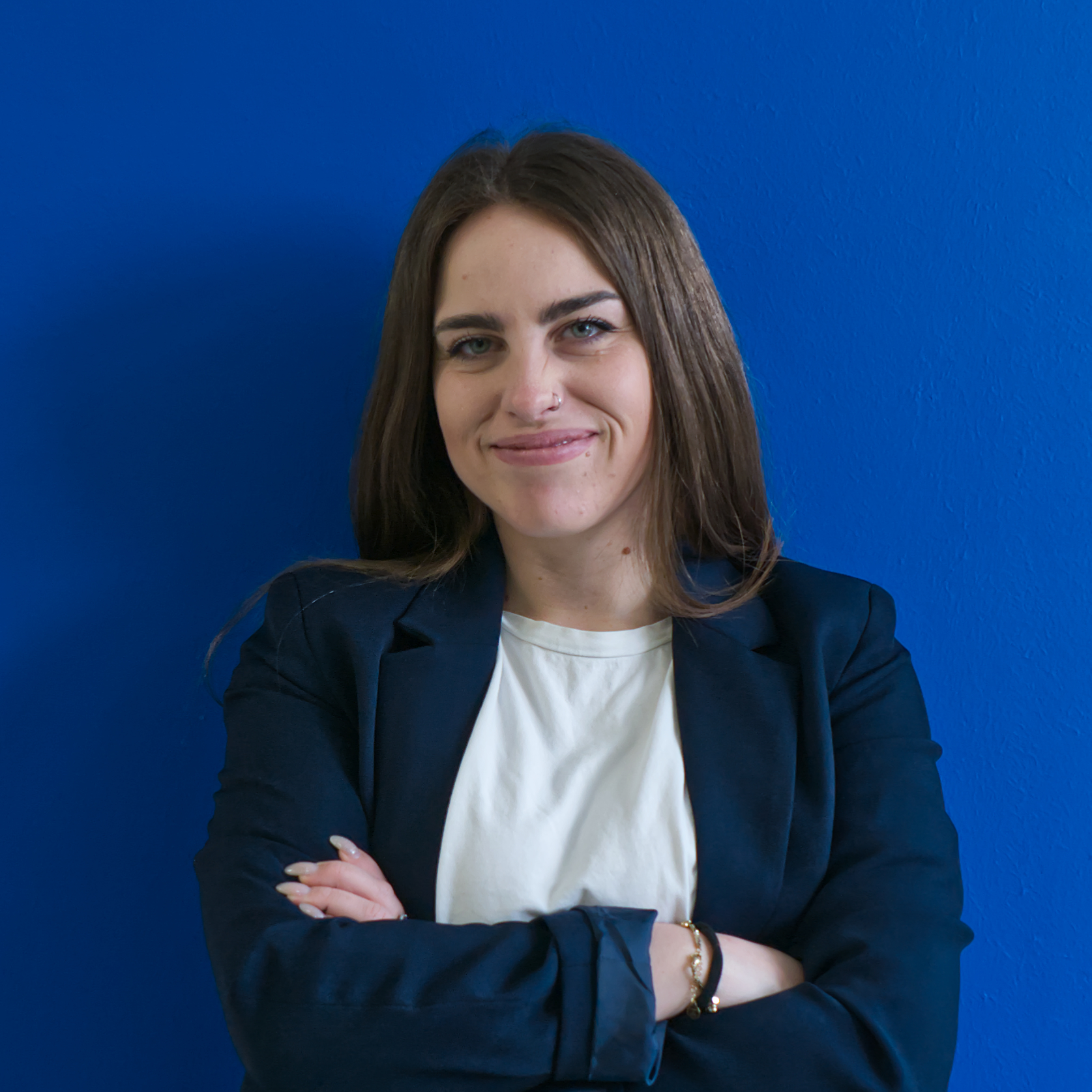 Una persona con lunghi capelli castani, che indossa un blazer blu navy e una camicia bianca, sta in piedi con sicurezza con le braccia incrociate. Sta sorridendo, incarnando lo spirito di 'Il team', con uno sfondo blu brillante dietro di loro.