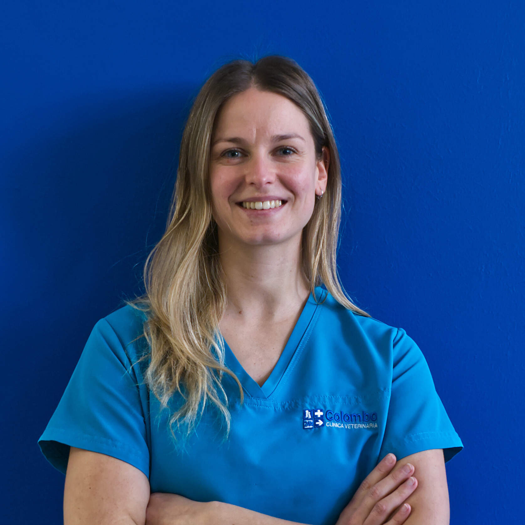 Una donna con indosso un camice veterinario blu è in piedi contro un muro blu brillante, sorride con sicurezza e tiene le braccia incrociate, incarnando lo spirito del team.
