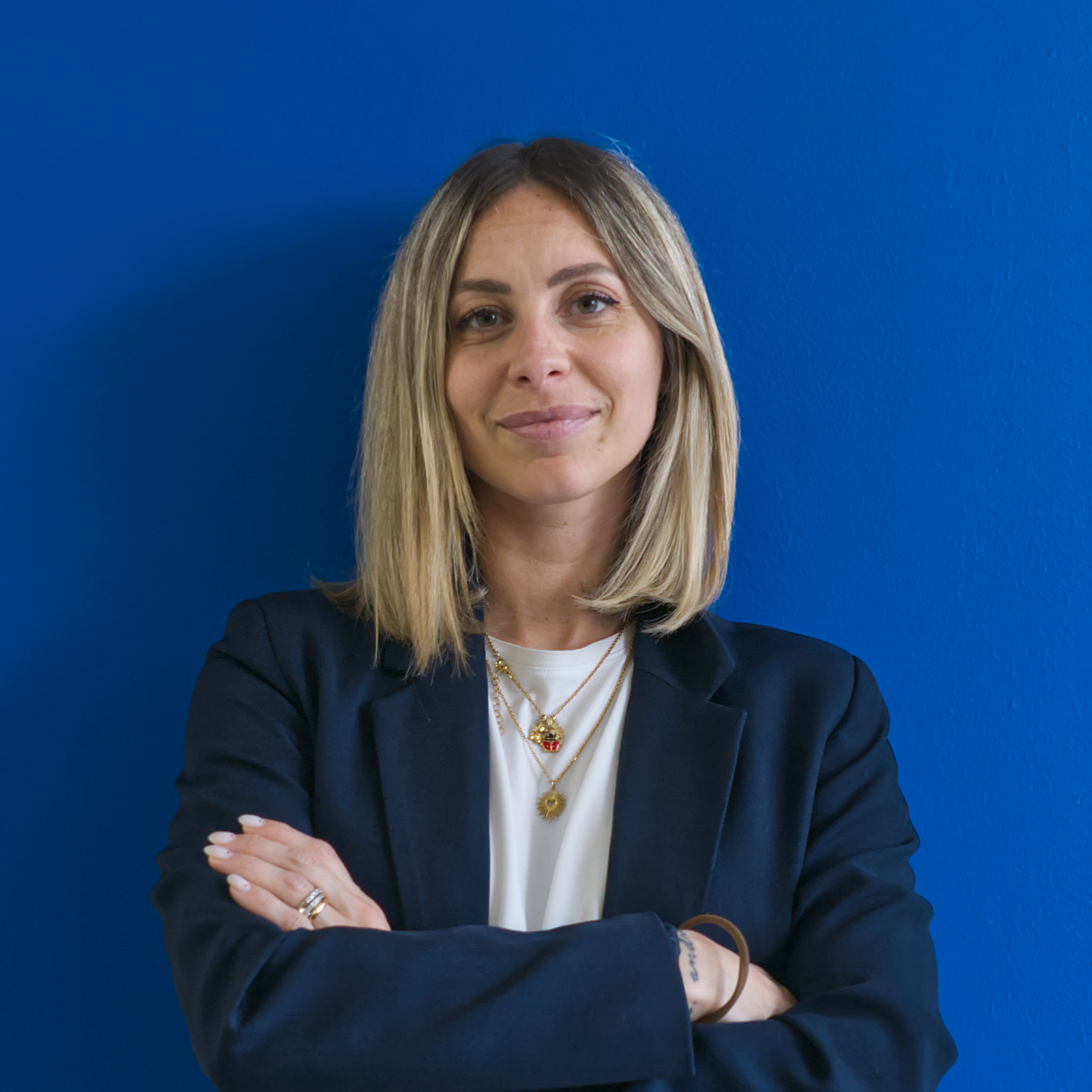 Una donna con i capelli biondi lunghi fino alle spalle è in piedi contro un muro blu brillante, sorridendo con sicurezza come se guidasse la squadra. Indossa un blazer scuro sopra un top bianco e accessoria con collane a strati, incarnando professionalità e stile.
