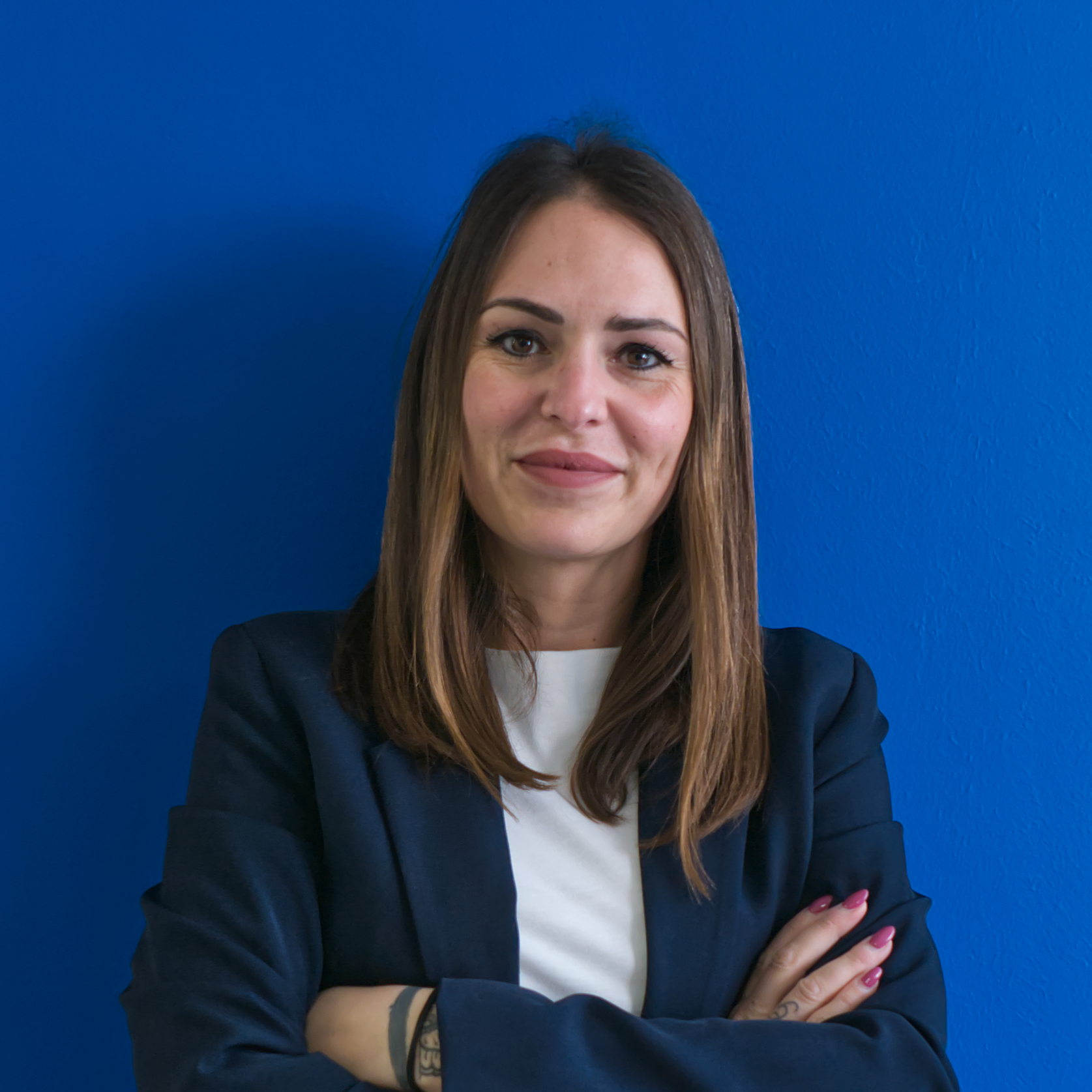 Una donna con lunghi capelli castani, giacca blu navy e top bianco, si erge con sicurezza su uno sfondo blu brillante, sorridendo e con le braccia incrociate, incarnando lo spirito della squadra.