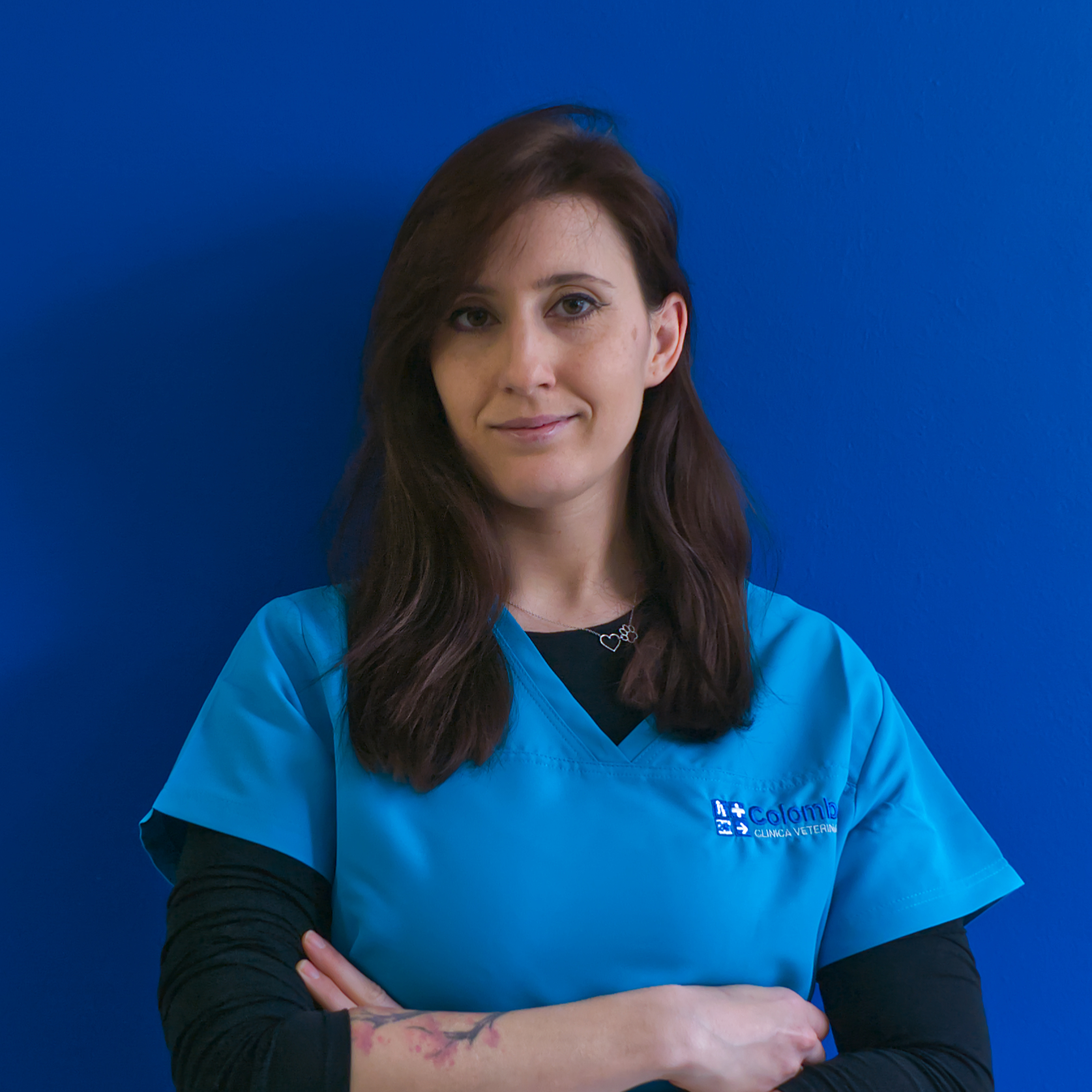 Una donna con lunghi capelli castani è in piedi su uno sfondo blu, indossa un camice medico blu e una maglietta nera sotto. Incrocia le braccia e sorride leggermente, trasudando sicurezza come membro integrante del team il.