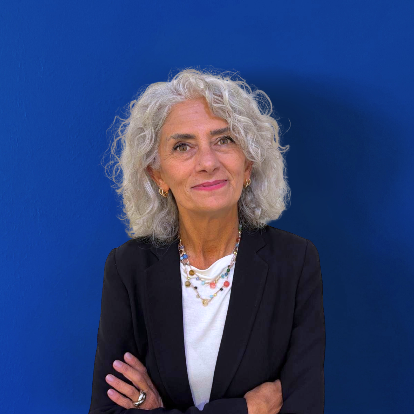 Una donna anziana con i capelli grigi e ricci è in piedi su uno sfondo blu uniforme. Indossa un blazer nero sopra un top bianco e completa il look con collane colorate. Il suo sorriso sicuro suggerisce che è la potenza dietro il team, con le braccia incrociate e pronta per la prossima sfida.