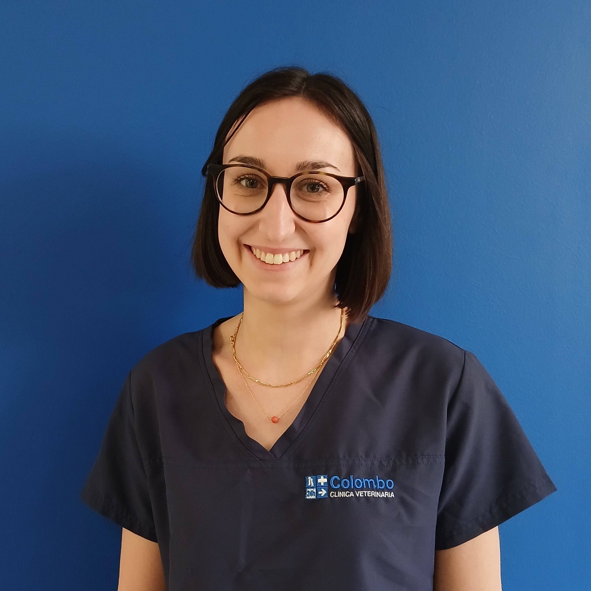 Una donna con capelli lisci e occhiali, che indossa un camice blu navy con la scritta Colombo Clínica Veterinaria, sta in piedi sorridendo davanti a una parete blu semplice come parte del team.